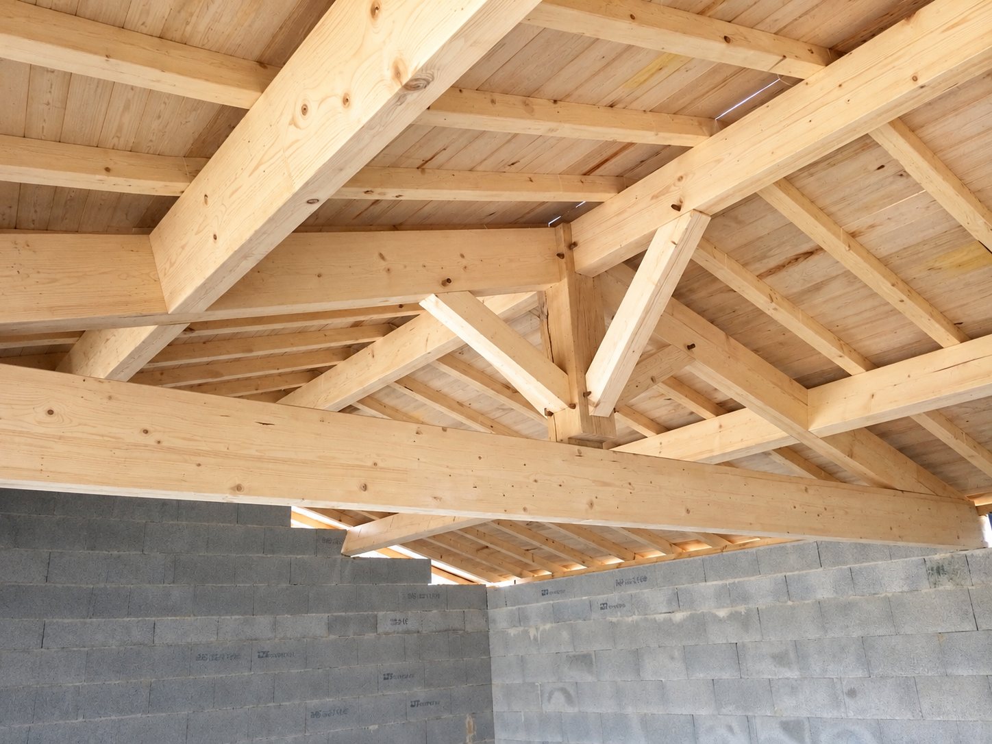Vue de l'intérieur d'un chantier montrant une charpente en bois clair posée sur des murs en blocs de béton gris.
