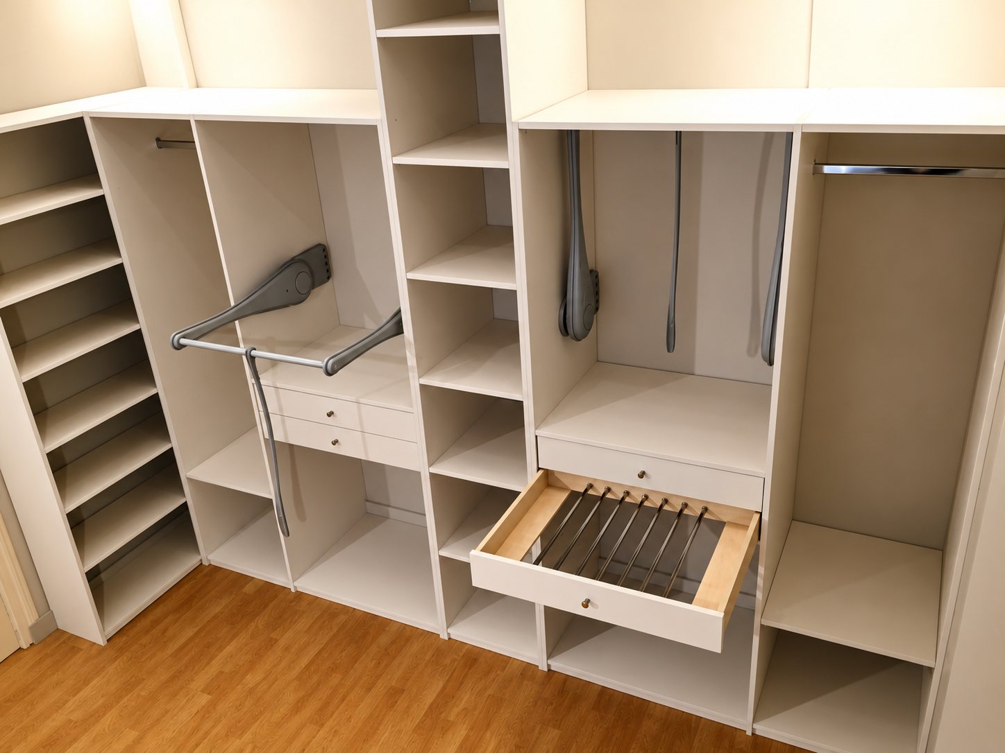 Dressing blanc vide installé dans une pièce à parquet, avec étagères, tiroirs, porte-pantalons ouvert et penderies coulissantes.
